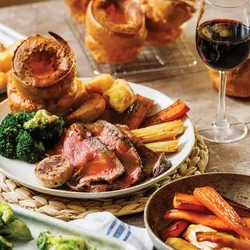 A beef roast dinner and a glass of red wine on a table surrounded by sides of vegetables.