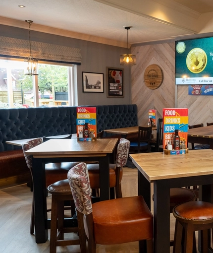 An interior restaurant seating area at The Heron; with wooden flooring; upholstered chairs, bar stools, and booth seating; a TV on the wall; and two illuminated dartboards at the far end of the room.