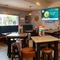An interior restaurant seating area at The Heron; with wooden flooring; upholstered chairs, bar stools, and booth seating; a TV on the wall; and two illuminated dartboards at the far end of the room.