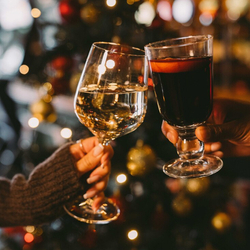 A close up view of two hands holding a glass of white wine and a glass of mulled wine, clinking them together in a cheers motion. A decorated and lit Christmas tree is visible in the background.