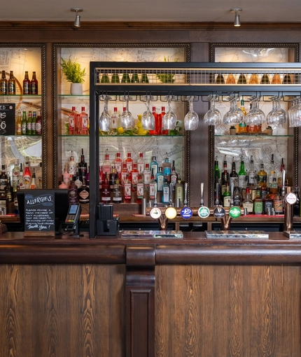 A close up view of the bar at The Man On The Moon, showing the beer taps, bottles of various spirits and wines on shelves, and glasses hanging in racks above the bar.