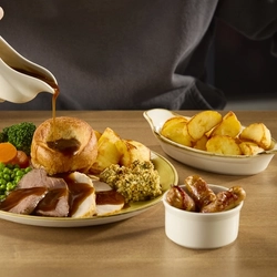 A Duo of Meat roast dinner at Hungry Horse venues, featuring slices of roast meat, roast potatoes, stuffing, Yorkshire pudding, peas, and vegetables, served on a plate. The plate sits on a wooden table next to a ramekin of pigs in blankets and a small roasting dish of roast potatoes, and a person sitting at the table is pouring gravy over the plate from a gravy boat.