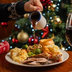 A close up view of a plate of carvery roast dinner sitting on a wooden table, while a hand pours gravy over the food from a jug. A glass of red wine also sits on the table, and a Christmas tree is visible in the background.
