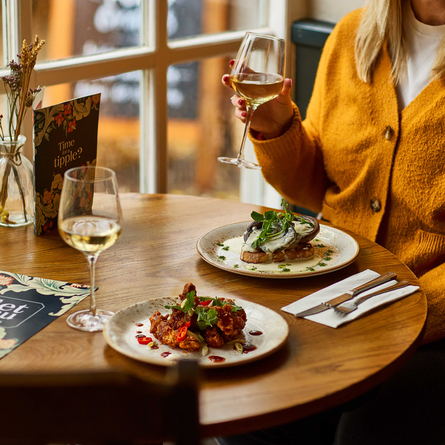 An image of a customer sat within the interior restaurant seating area enjoying a glass of white wine with 2 Starter dishes and a glass of white wine sat on the table from the Set menu at a Chef & Brewer venue.