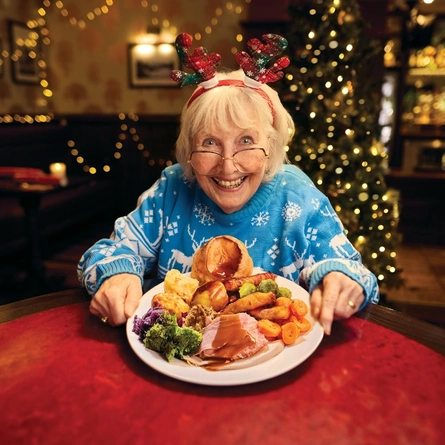 A roast dinner featuring roast meat, Yorkshire pudding, vegetables and gravy, served on a plate which sits on a table. A woman is sitting at the table, holding the plate with both hands and smiling at the camera.