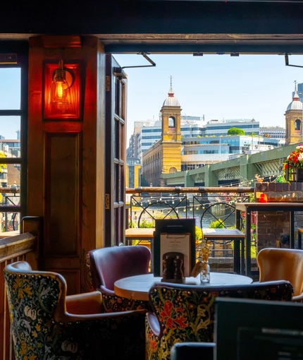 An interior restaurant seating area at The Anchor Bankside in Southwark; with upholstered chairs, wall lights on the wood panelled walls, and open doors leading out into the beer garden overlooking the River Thames.