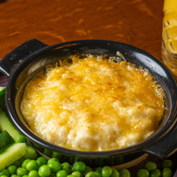 A close up view from above of a children's Mac & Cheese in a small roasting dish, served on a plate with peas and vegetable sticks. The plate sits on a wooden restaurant table along with a glass of orange juice.