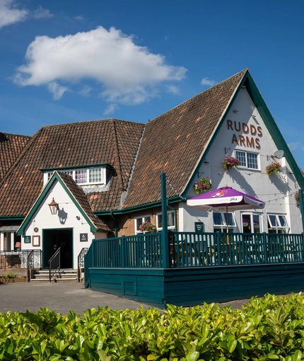 The exterior facade, signage, and seating area of the Rudds Arms in Marton-In-Cleveland; with an accessibility ramp leading up to the door; and picnic tables, shade umbrellas, and string lights on a raised deck area.