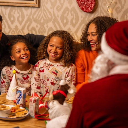 A festive breakfast table in a Christmas-themed restaurant booth. Two children wearing Christmas-print pyjamas sit beside an adult in an orange jumper, while a person dressed as Santa Claus is seated in the foreground with their back to the camera. One child wears a green-and-red elf hat. The table is set with breakfast plates, juice cartons, a party hat, Christmas colouring activities, and festive decorations. Warm lighting, patterned wallpaper and seasonal décor create a cosy Christmas setting.