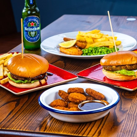A bowl of chicken nuggets with dipping sauce, plates of burgers, chips and onion rings, glasses of drinks, and a bottle of Heineken low alcohol beer, sitting on a table.