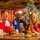 Three football fans sit around a wooden table in a sports pub decorated for Christmas, holding glasses of beer beside a frosted Christmas tree. One person wears a Santa hat and another wears reindeer antlers, while burgers topped with onion rings and pickle skewers, chips and dipping sauces are served on the table. Live football is shown on large screens in the background, creating a festive Boxing Day matchday atmosphere.