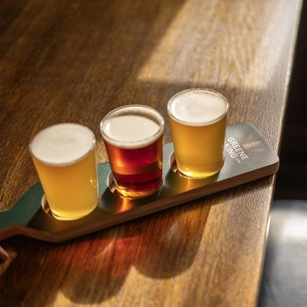 Three half pint glasses of an ale, a craft beer, and a cider, sitting in a paddle shaped drinks holder on a wooden table inside a pub.