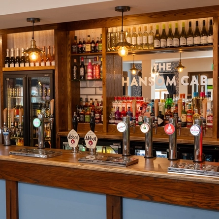 A close up view of the bar at The Hansom Cab in Hinckley, with bottles of wine and alcoholic spirits on wooden shelves behind the counter.