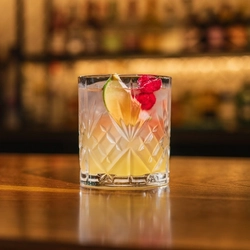 A close up view of a Pineapple Margarita served in a whiskey glass with ice, dried raspberries, and a lime wedge. The glass stands on the bar inside a pub.