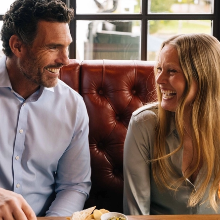 Four people seated at a wooden table which is holding sharing boards and plates of pub food, including halved Scotch eggs, pickles, fried bites, bread with dips, and loaded nachos, with glasses of soft drinks and a pint of Hazy Day. Only two people are showing in the image.