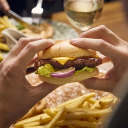 A close up view of a burger being held in two hands, above a table laden with plates of food and glasses of wine.