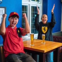 Two people sit at a wooden high-top table in a pub, enthusiastically celebrating while watching a sporting event. One person wears a red Ferrari cap and matching red shirt, raising both arms in triumph. The other person, dressed in a black Ferrari hoodie, stands slightly behind the table with their arms lifted in applause. Two tall pints of beer sit on the table in front of them. The room features blue and beige walls, framed artwork, and a mix of pub-style seating, creating a lively and supportive atmosphere for watching Formula One.