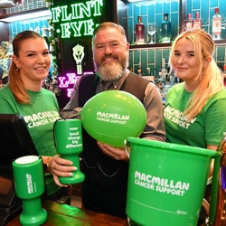 Three people with Macmillan T shirts, balloons and collection buckets behind a bar.