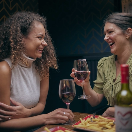 Two women laughing together and holding glasses of red wine, sitting at a wooden table laden with plates of burgers and chips.