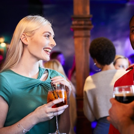 Two people inside a crowded bar area, smiling and holding drinks.