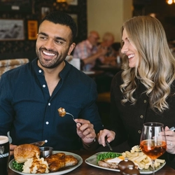 Two people seated at a pub table eating sausages with mash, peas and Yorkshire pudding, and gammon with egg, peas and chips, with a pint of Guinness and a glass of rosé.