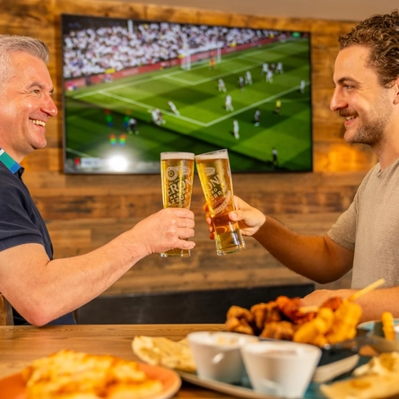 A son and Father holding a pint of Peroni each into a 'cheers' position, food is blurred on the table in-front of them and sports are on the TV blurred in the background at a Flaming Grill venue on Fathers day.