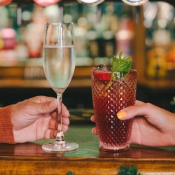 A glass of champagne and a cocktail served in a highball glass and garnished with mint, sitting on a bar inside a pub. A hand holds each glass and a Christmas garland is attached to the bar.