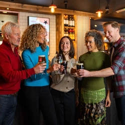 An image of a group of people enjoying drinks and celebrating with a cheers, stood at the bar within a Flaming Grill venue.