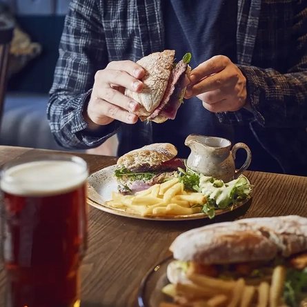 A roast beef sandwich on a table being picked up and eaten, with another sandwich and a pint of Greene King IPA in front of it.