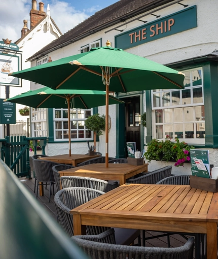 The exterior facade and seating area at the front of The Ship; with shade umbrellas on some of the wooden tables, and flower baskets hanging on the white walls of the pub.