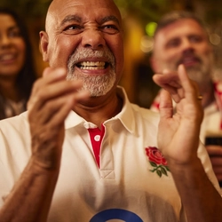 A person wearing a white rugby-style shirt with a red rose emblem stands in a warmly lit pub, clapping enthusiastically. They are surrounded by other fans, including someone behind them wearing a red and white scarf and holding a pint of stout. The background shows softly blurred pub lighting and glassware, creating a lively atmosphere associated with watching a live sports match.