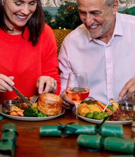 Three people seated around a table in a festively decorated pub, enjoying a traditional Christmas meal. Plates containing roast dinners with Yorkshire puddings, vegetables and roast potatoes are served alongside a pint of beer, glasses of red and rosé wine, and green Christmas crackers. Seasonal garlands, framed artwork, patterned wallpaper and warm decorative lighting create a festive dining atmosphere.