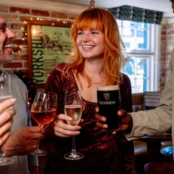 A group gathered in a pub raising drinks during a toast. In the centre, a person in a red patterned dress holds a glass of sparkling wine while others hold a rosé wine, sparkling wine and a pint of stout. The venue features exposed brick walls, framed artwork, booth seating and warm decorative lighting.