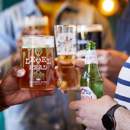 A close up view of a hand holding a glass of Level Head IPA. Other people holding glasses of beer, stout, and a bottle of Peroni 0.0% alcohol beer are visible in the background.