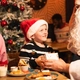 A child wearing a Santa hat sits at a table in a pub decorated for Christmas, receiving a wrapped gift from a person dressed as Santa Claus. An adult sits beside the child, while a snow-covered Christmas tree with red baubles stands in the background. Breakfast plates with milk, fruit and cooked food are visible on the table, creating a festive family dining scene.