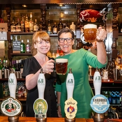 An image showing 2 people stood behind the interior bar at The Two Chairmen holding up drinks in celebration.
