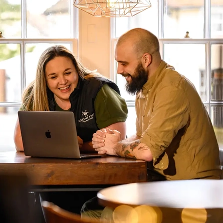 A Woman and Man sat at a table looking at a laptop and smiling