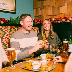 Three people sit in a festive pub booth decorated with Christmas garlands and red baubles. A wooden table is spread with sharing dishes including pigs in blankets, roast potatoes, dipping sauces and burgers, alongside glasses of lager, stout and red wine. One person takes a large bite from a stacked burger while the others enjoy food and drinks in a cosy Christmas pub setting.