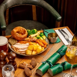 A plated turkey dinner on a festive decorated wooden table with a tankard of beer, a glass and bottle of sparkling wine and a stemmed glass containing a spirit cocktail.