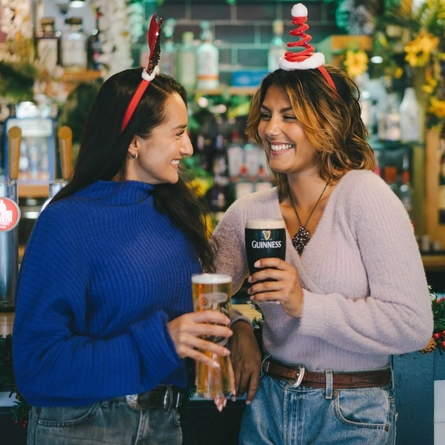 An image of 2 friends enjoying drinks stood at the interior bar within a Greene King venue.