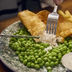 A close up view of a plate of Fish, Chips and Peas, served with a small pot of sauce. The plate sits on a wooden table and a person is cutting up the fish with their knife and fork.