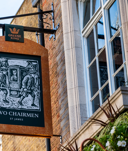 A close up view of the pub sign hanging on the exterior wall of the Two Chairmen. The sign features a black and white woodcut-style illustration of two men using poles to carry a sedan chair with a woman inside, above the words "Two Chairmen, St James". 
