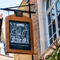 A close up view of the pub sign hanging on the exterior wall of the Two Chairmen. The sign features a black and white woodcut-style illustration of two men using poles to carry a sedan chair with a woman inside, above the words "Two Chairmen, St James". 