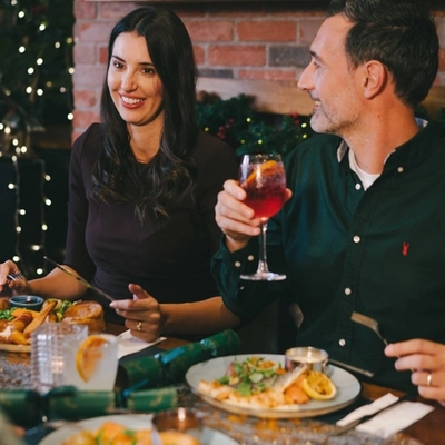 An image focusing on 2 people enjoying various main dishes and a selection of drinks while sat within the interior restaurant seating area.
