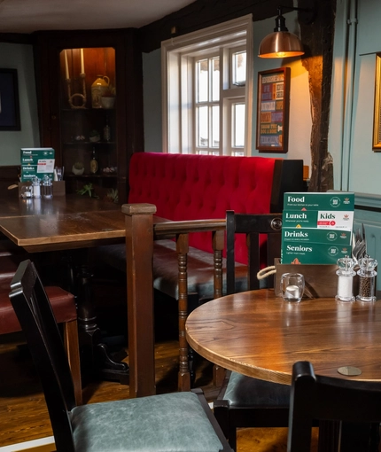 An interior restaurant seating area at The Ship; with an upholstered booth seat against the window, framed artwork on the walls, and wooden beams on the walls and ceiling.
