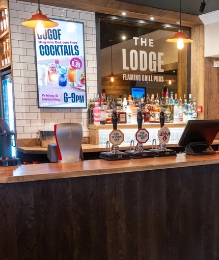 The bar inside The Lodge in Alvaston, with TVs on the wall, and bottles of wine and alcoholic spirits on wooden shelves behind the counter.