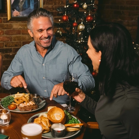 Three people sitting at a restaurant table with Christmas crackers and plates of food in front of them. One person is holding a glass of red wine, one person is holding a glass of white wine, and the third person is cutting up their food with their knife and fork.