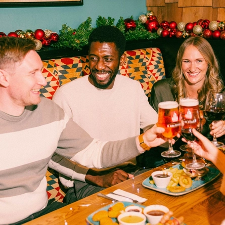 Four people sit together in a festive pub booth decorated with Christmas garlands and red baubles. The group raises glasses of lager and red wine in a toast over a wooden table filled with pub food, including burgers, onion rings, pigs in blankets and dipping sauces, creating a warm and social Christmas dining scene.