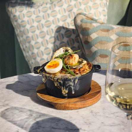 An advertising image showing a Fish Pie dish served in a roasting dish sat on a table with a glass of white wine within the interior of The White Horses.