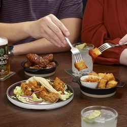 A close-up of a table with small plates of food. Two people are reaching in with forks, and drinks such as beer and cocktails are placed around the table.